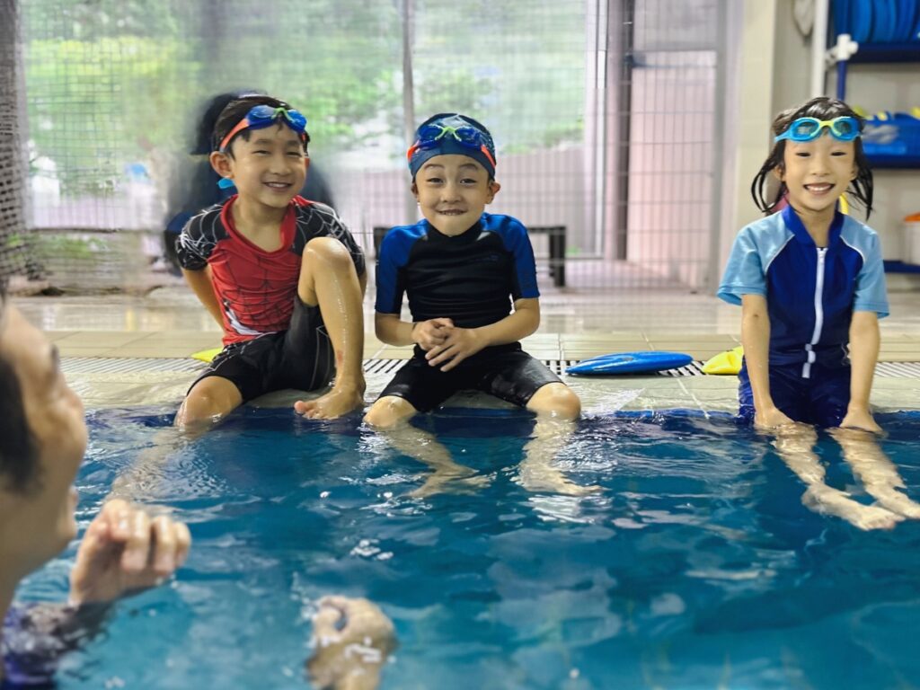 Three young children wearing swimwear and goggles sit on the edge of a swimming pool, smiling and enjoying themselves, while an adult instructor in the water engages with them. Behind them, various swim gear and clothes are visible hanging or stored on shelves.