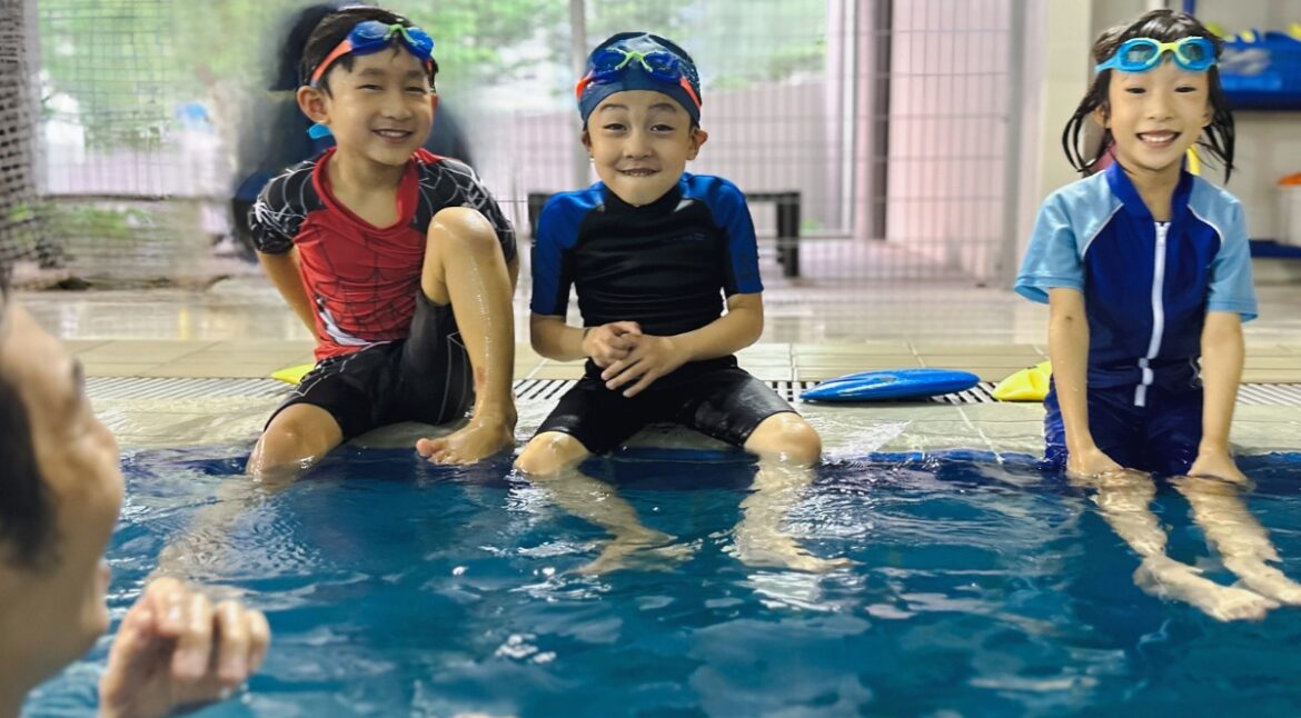 Three young children wearing swimwear and goggles sit on the edge of a swimming pool, smiling and enjoying themselves, while an adult instructor in the water engages with them. Behind them, various swim gear and clothes are visible hanging or stored on shelves.