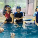 Three young children wearing swimwear and goggles sit on the edge of a swimming pool, smiling and enjoying themselves, while an adult instructor in the water engages with them. Behind them, various swim gear and clothes are visible hanging or stored on shelves.