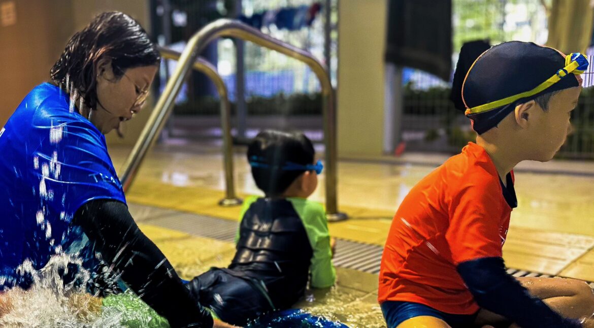 A swim instructor in a blue shirt sits by the pool, supporting two young children as they practice water entry and play at the pool's edge. One child in a green swimsuit and another in an orange shirt sit beside her, ready to begin their lesson.