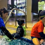 A swim instructor in a blue shirt sits by the pool, supporting two young children as they practice water entry and play at the pool's edge. One child in a green swimsuit and another in an orange shirt sit beside her, ready to begin their lesson.