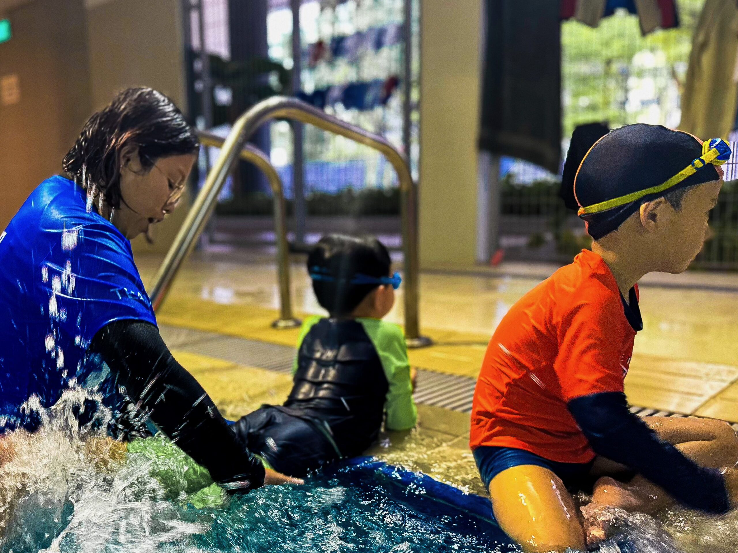 A swim instructor in a blue shirt sits by the pool, supporting two young children as they practice water entry and play at the pool's edge. One child in a green swimsuit and another in an orange shirt sit beside her, ready to begin their lesson.