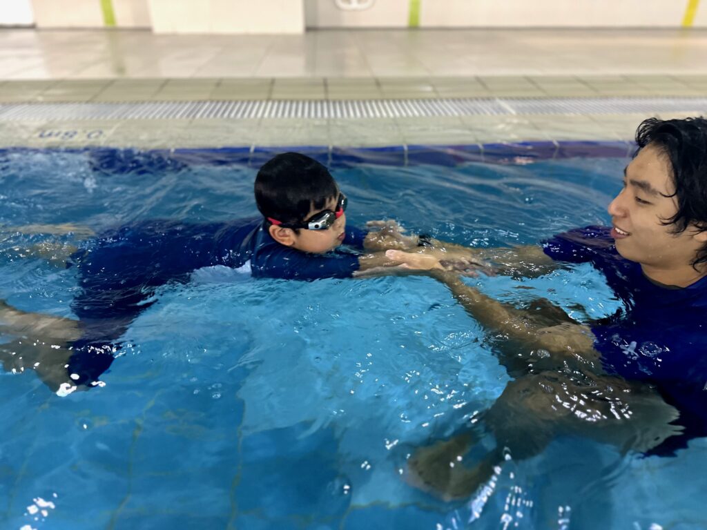 A young child practices swimming with the guidance of an instructor in the pool. The instructor holds the child’s hands for support as they float on the water, helping the child build confidence and improve technique.