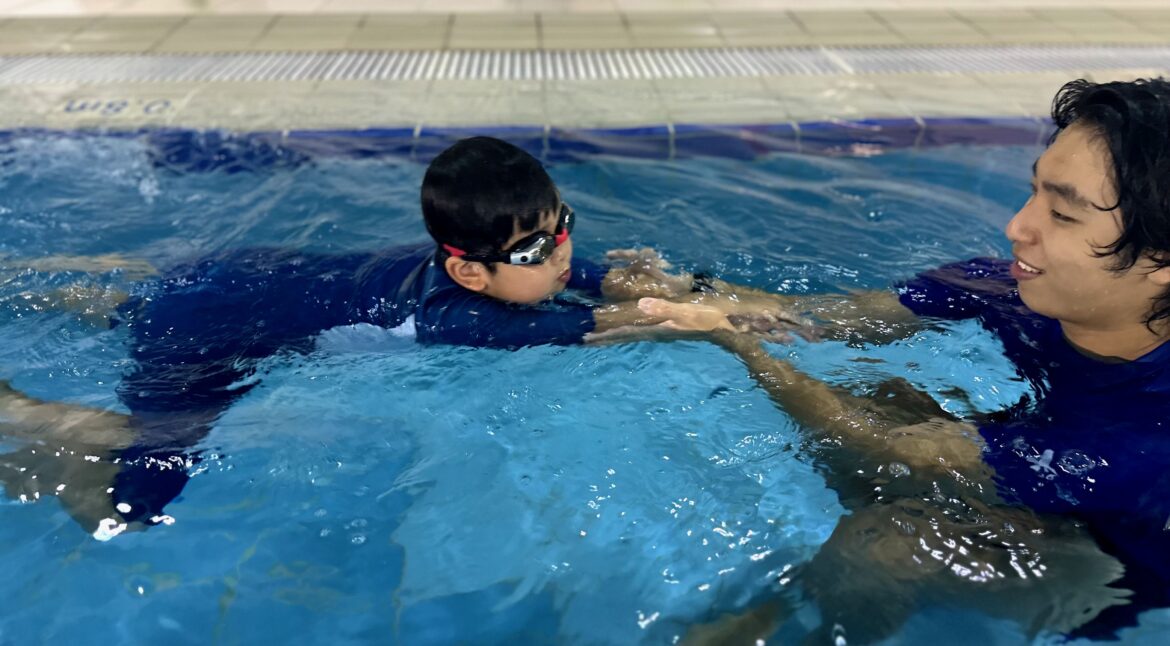 A young child practices swimming with the guidance of an instructor in the pool. The instructor holds the child’s hands for support as they float on the water, helping the child build confidence and improve technique.