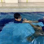 A young child practices swimming with the guidance of an instructor in the pool. The instructor holds the child’s hands for support as they float on the water, helping the child build confidence and improve technique.