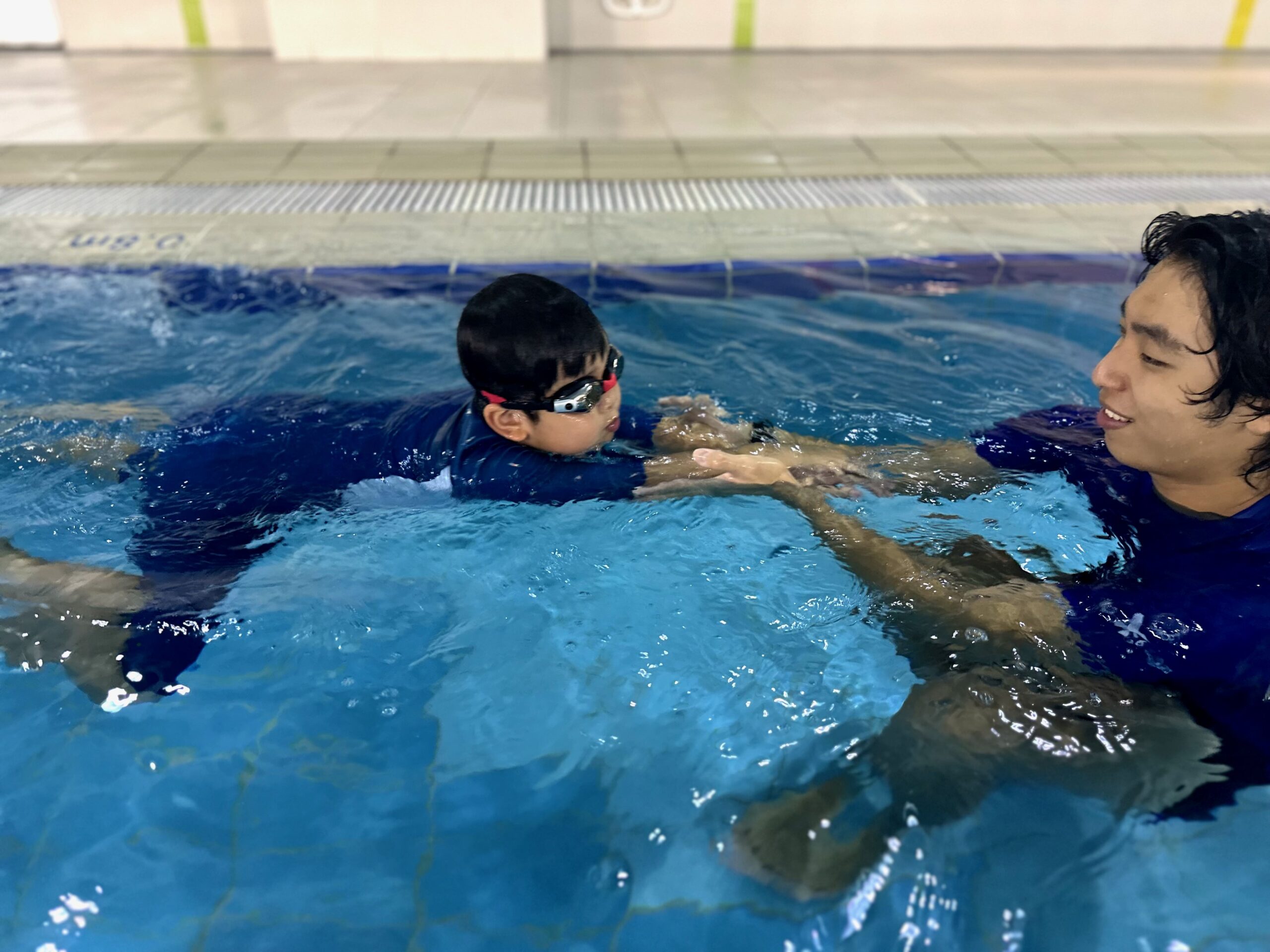 A young child practices swimming with the guidance of an instructor in the pool. The instructor holds the child’s hands for support as they float on the water, helping the child build confidence and improve technique.