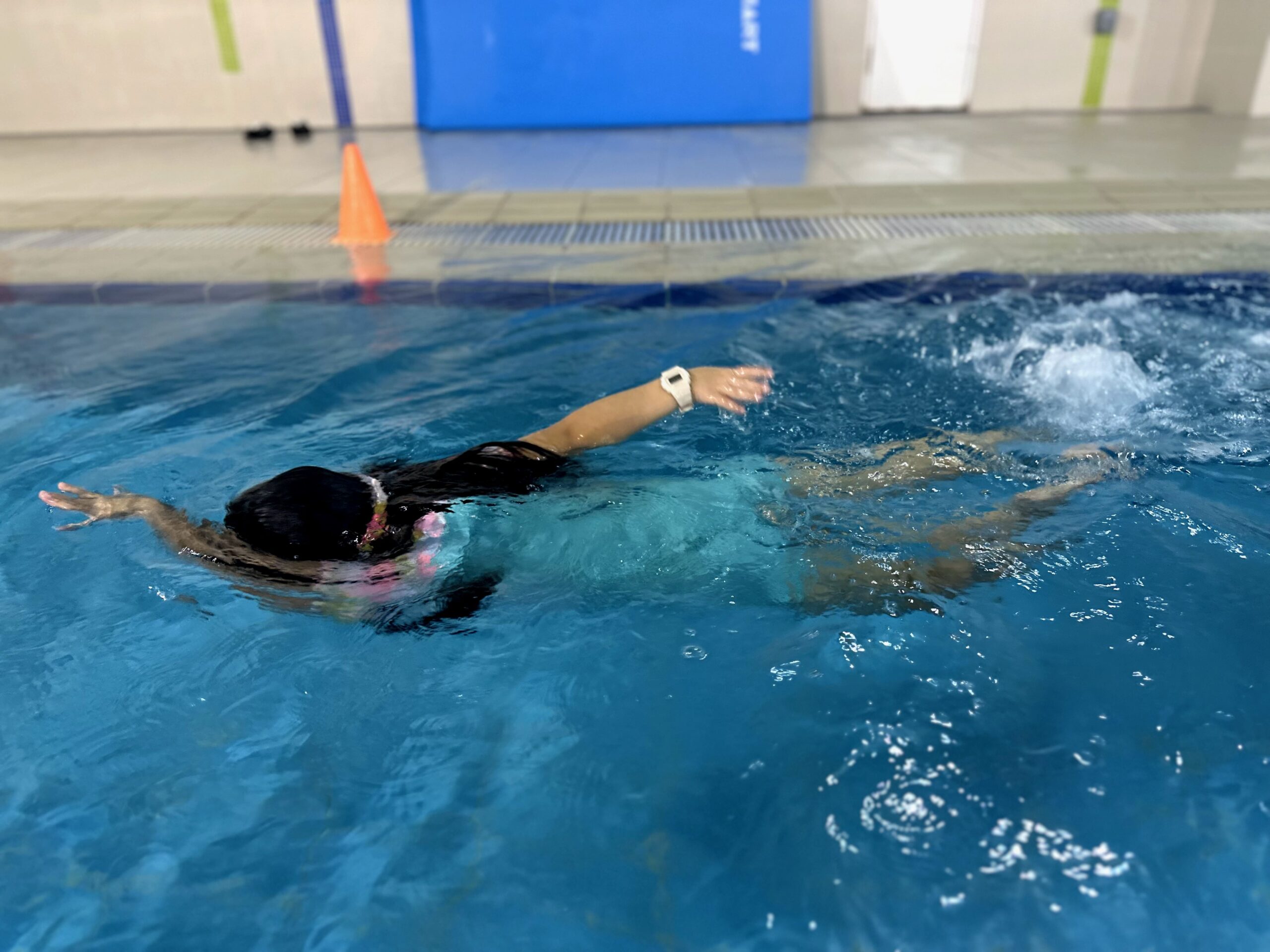 A young swimmer in a pool performs a freestyle stroke with one arm extended forward. The swimmer’s head is turned to the side, partially submerged in the water, as they propel forward, creating a splash behind them.