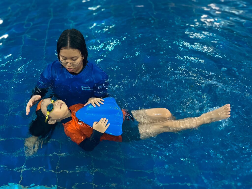 A swimming instructor in a blue shirt is assisting a young child wearing orange and black swim gear and goggles in the water. The child is holding a blue kickboard and floating on their back, with the instructor supporting their head and guiding them.