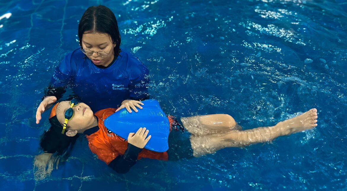 A swimming instructor in a blue shirt is assisting a young child wearing orange and black swim gear and goggles in the water. The child is holding a blue kickboard and floating on their back, with the instructor supporting their head and guiding them.