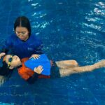 A swimming instructor in a blue shirt is assisting a young child wearing orange and black swim gear and goggles in the water. The child is holding a blue kickboard and floating on their back, with the instructor supporting their head and guiding them.