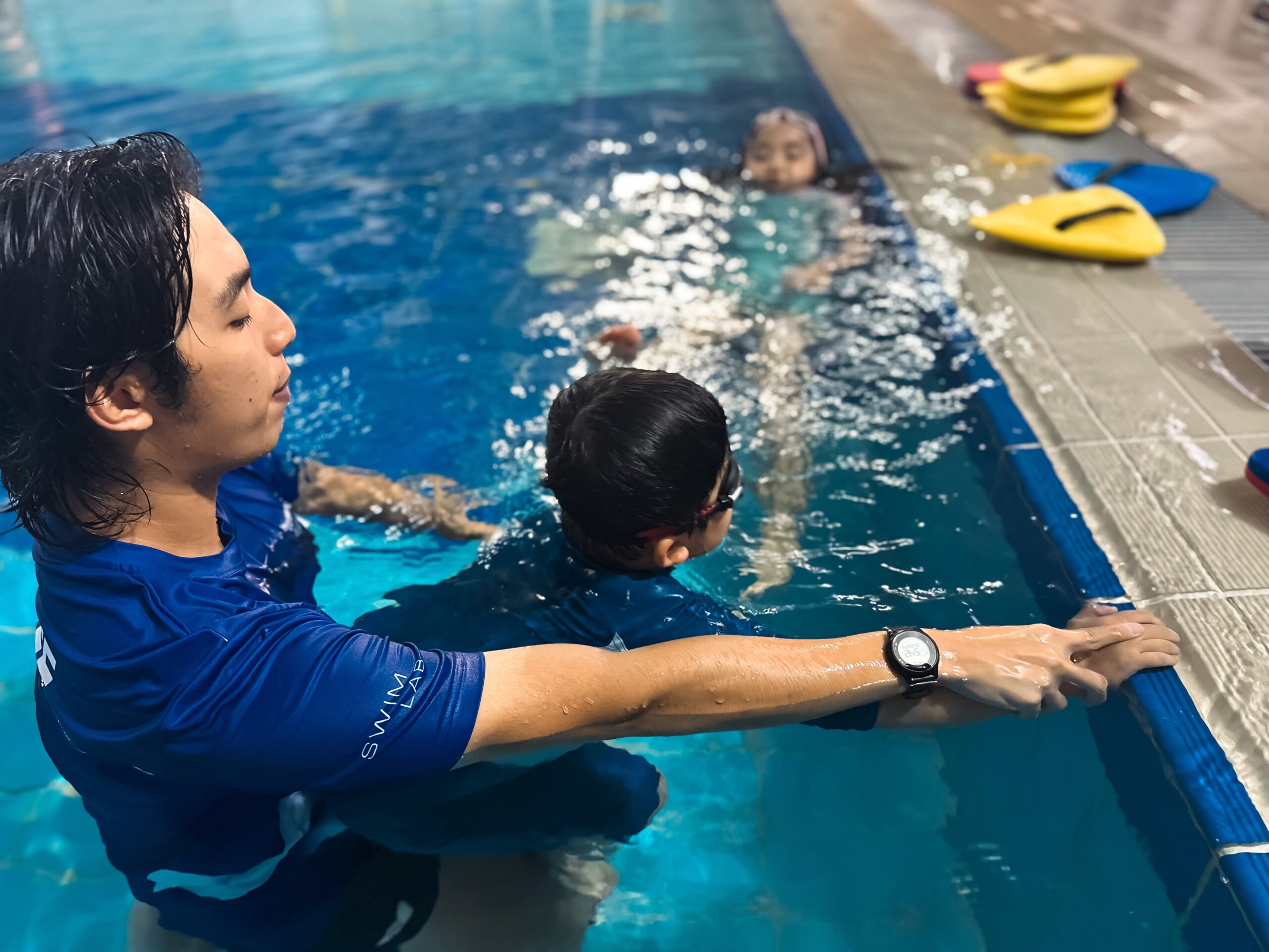 A swimming instructor in a blue shirt is holding a young child in a pool, helping them practice swimming techniques near the pool edge. The child is wearing goggles and is gripping the pool ledge for support, while another child is swimming nearby. Several colorful kickboards are visible on the pool deck.