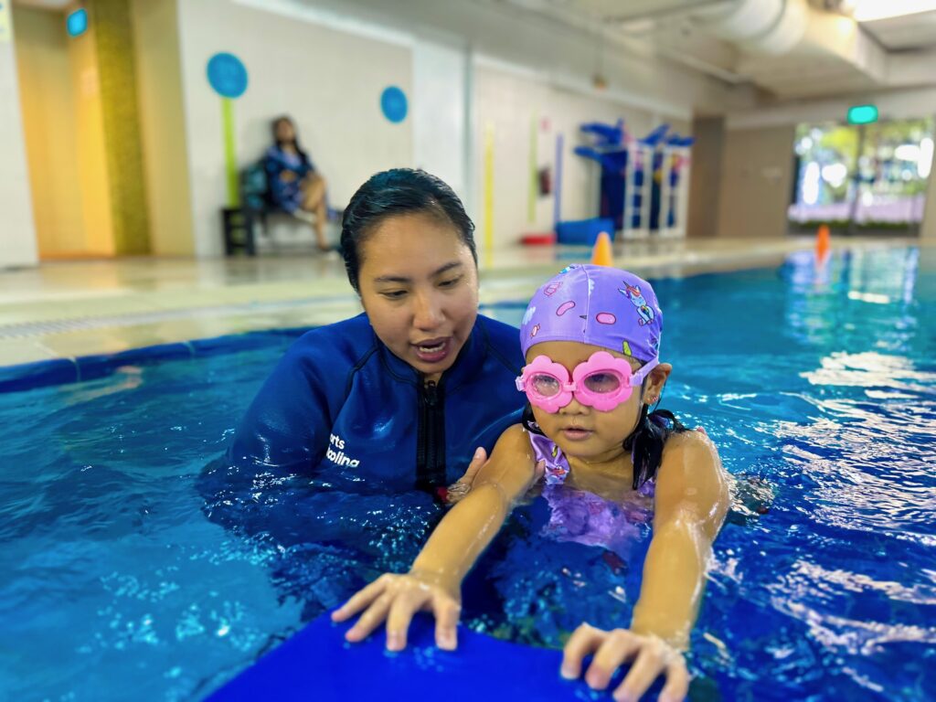 A young child wearing a purple swim cap with colorful designs and pink heart-shaped goggles is learning to swim in a pool. They hold onto a blue kickboard with guidance from their instructor, who is wearing a blue wetsuit and providing close support.