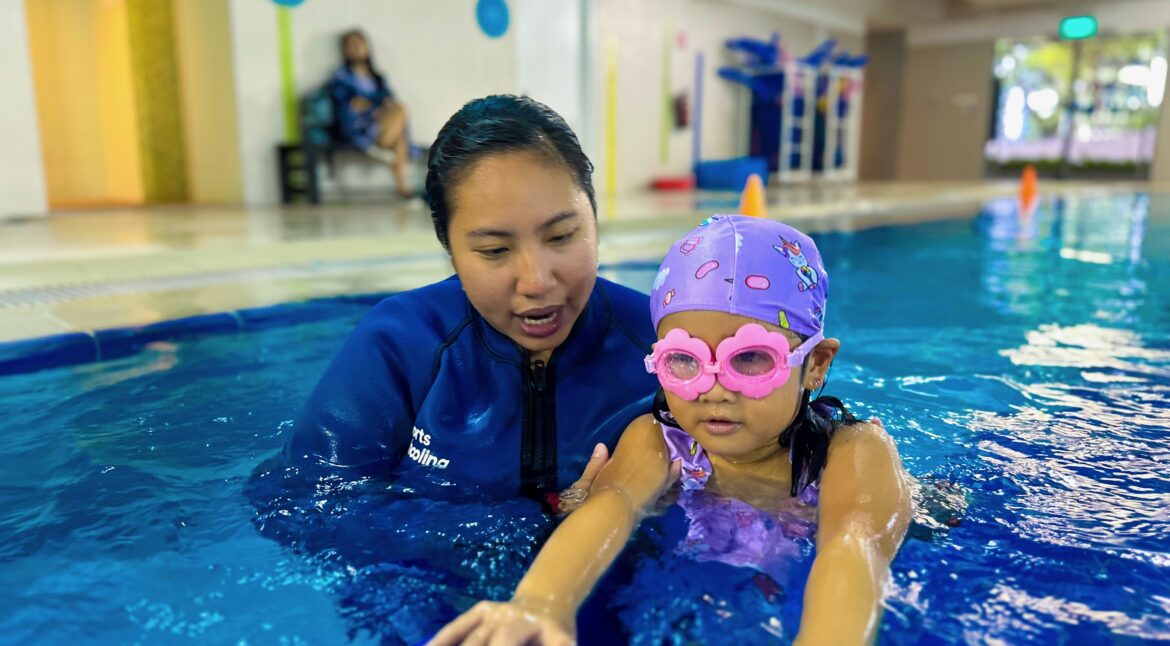 A young child wearing a purple swim cap with colorful designs and pink heart-shaped goggles is learning to swim in a pool. They hold onto a blue kickboard with guidance from their instructor, who is wearing a blue wetsuit and providing close support.