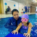 A young child wearing a purple swim cap with colorful designs and pink heart-shaped goggles is learning to swim in a pool. They hold onto a blue kickboard with guidance from their instructor, who is wearing a blue wetsuit and providing close support.